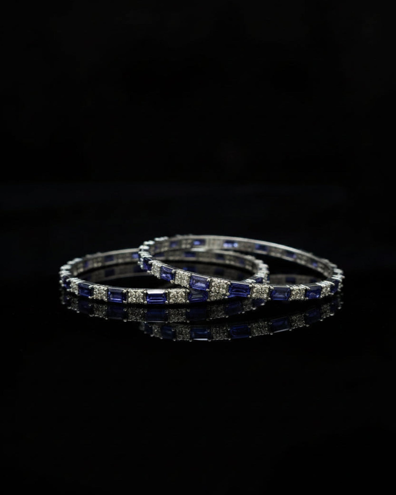 A pair of silver-toned bangles decorated with alternating deep blue rectangular gemstones and clear round stones, displayed on a reflective black surface against a black background.
