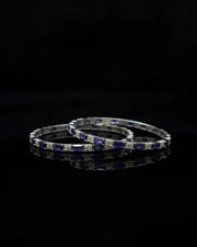 A pair of silver-toned bangles decorated with alternating deep blue rectangular gemstones and clear round stones, displayed on a reflective black surface against a black background.