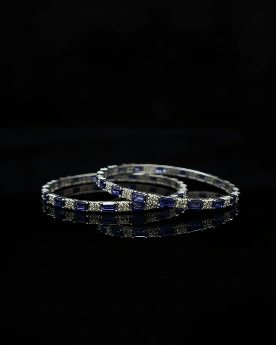 A pair of silver-toned bangles decorated with alternating deep blue rectangular gemstones and clear round stones, displayed on a reflective black surface against a black background.