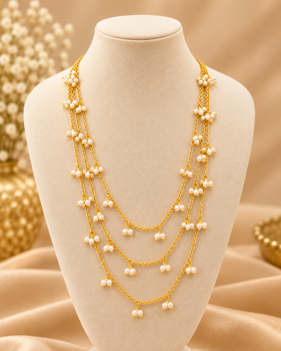 A multi-strand gold chain necklace adorned with small white pearl-like floral clusters, displayed on a black jewelry bust against a warm beige satin background.