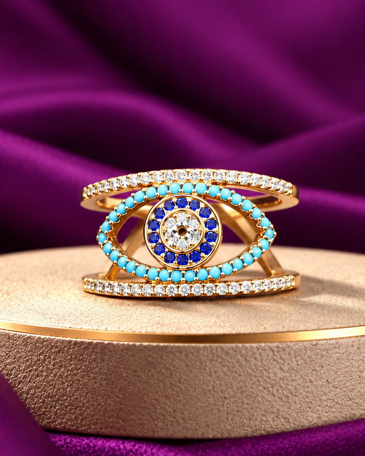 A gold evil eye ring with turquoise and blue accents, set with small clear stones, displayed on a round beige pedestal against a draped purple satin background.