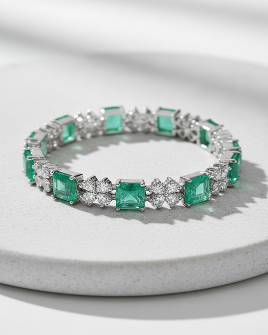 A silver-toned tennis bracelet featuring square-cut emerald-cut green gemstones and round brilliant-cut clear stones, displayed on a light gray textured circular pedestal.