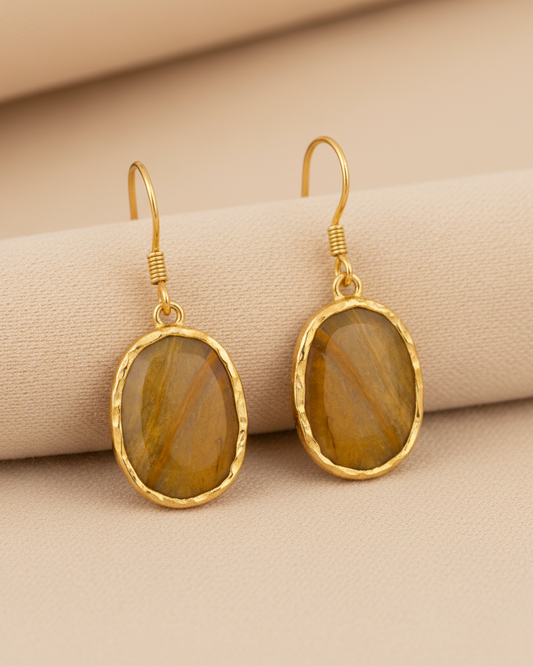 A pair of gold drop earrings with oval-shaped brown tiger's eye stones, displayed on a beige textured fabric background.