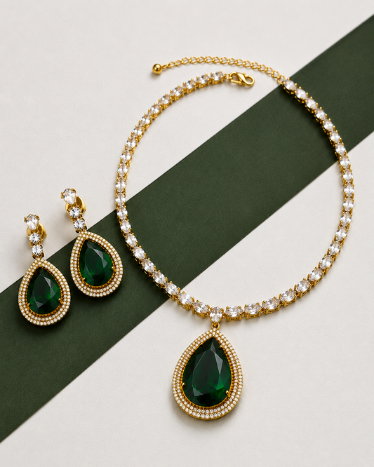 Gold jewelry set featuring a teardrop pendant necklace and matching drop earrings, with deep green teardrop gemstones and clear rhinestone borders, displayed on a diagonal dark green ribbon over a light beige background.