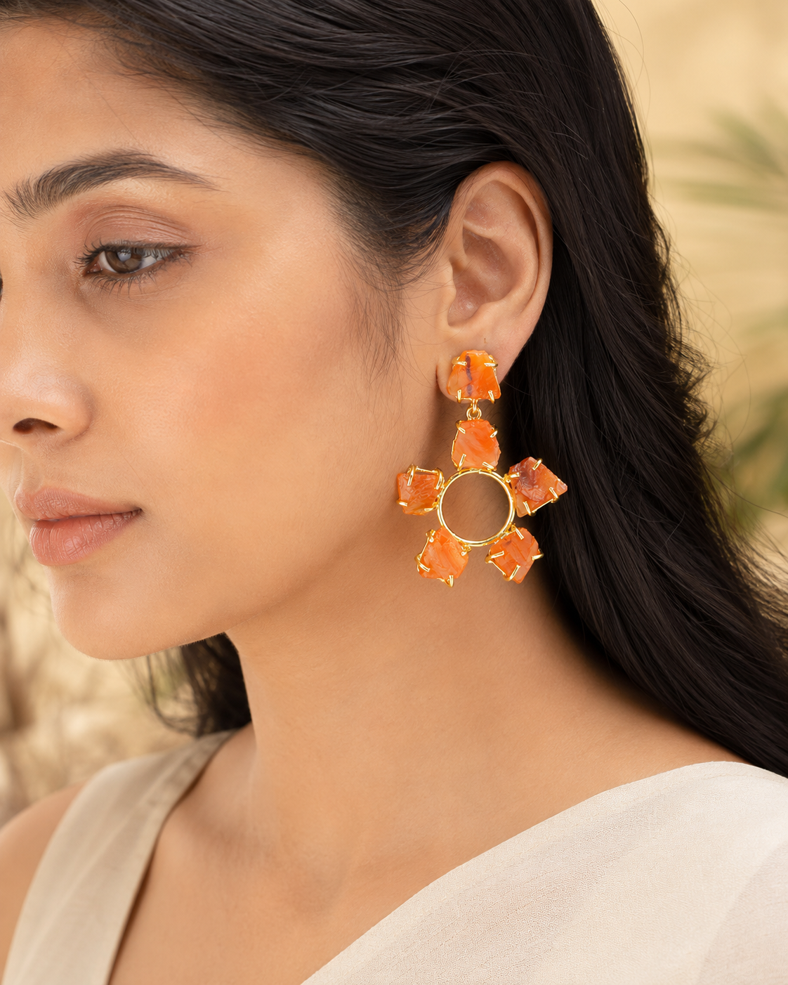 A close-up of a woman wearing a large gold-toned dangle earring with six orange stone-like petals arranged in a flower shape.