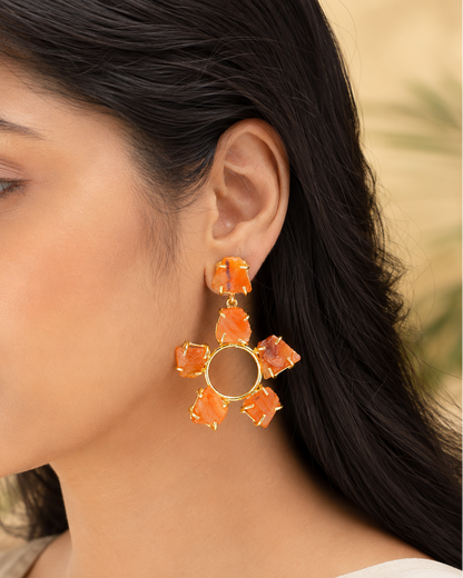 A close-up of a woman wearing a large gold-toned dangle earring with six orange stone-like petals arranged in a flower shape.
