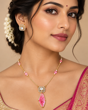 A close-up portrait of a woman wearing a gold-toned necklace with a large pink teardrop pendant and pink bead accents, along with matching earrings, against a warm beige background.