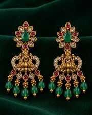 A pair of ornate gold-toned Indian-style dangle earrings with green teardrop emeralds, pinkish-red round rubies, clear stones, and hanging green beads, displayed on a dark green velvet background.