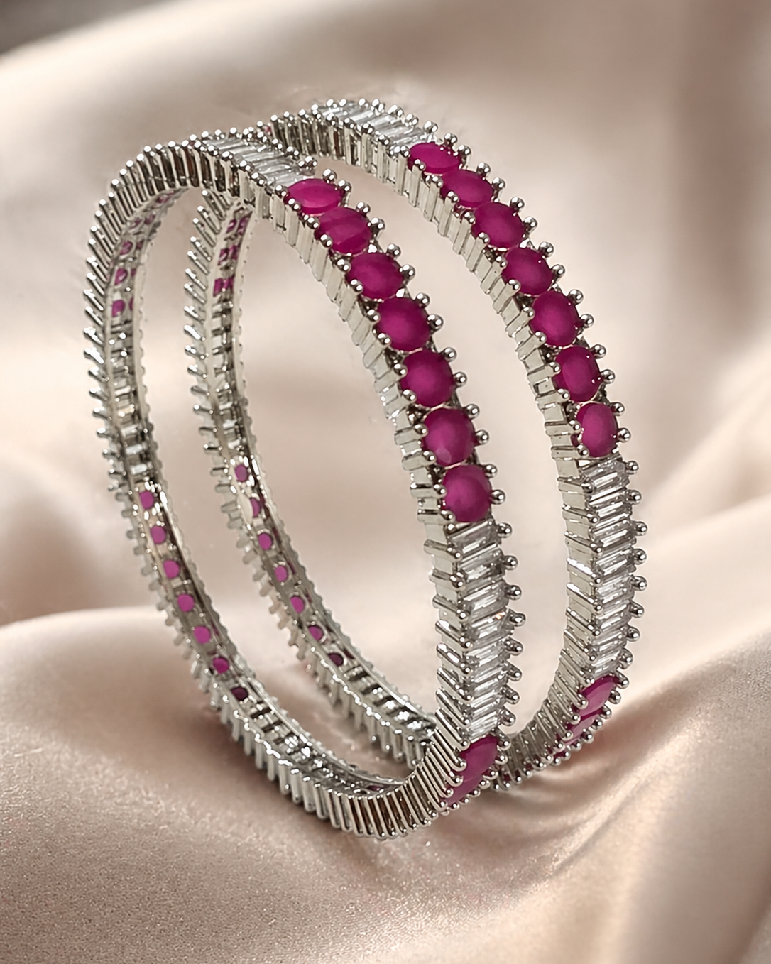 A pair of silver-toned bangles with rows of bright pink oval gemstones, displayed on a draped beige satin fabric background.