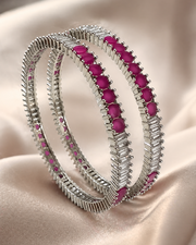A pair of silver-toned bangles with rows of bright pink oval gemstones, displayed on a draped beige satin fabric background.