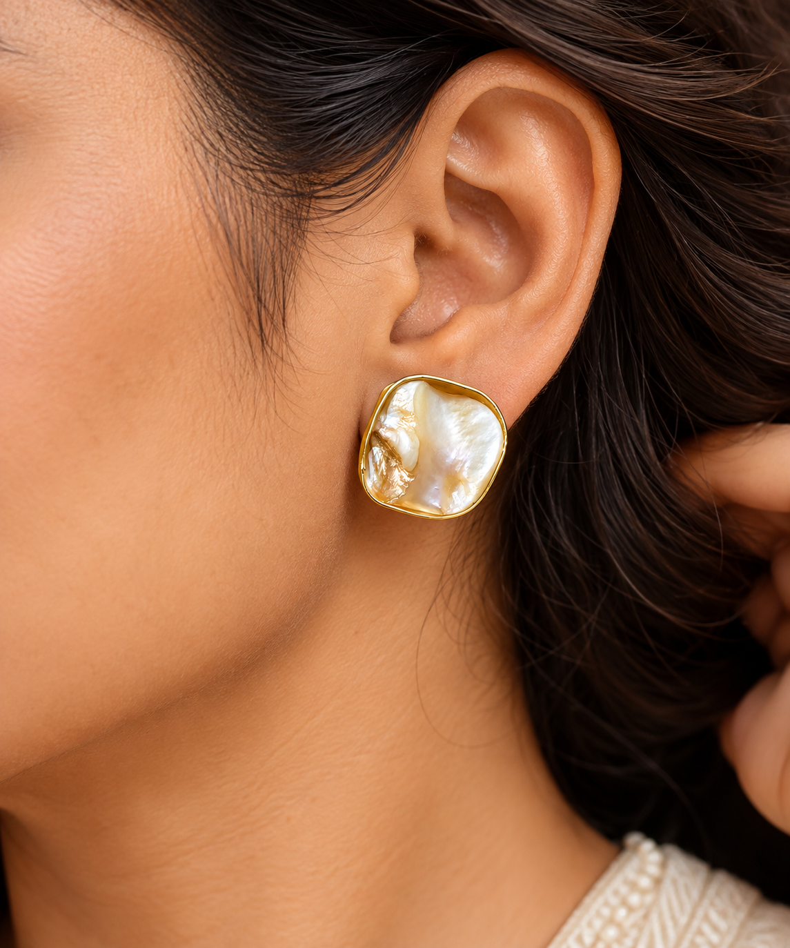A close-up of a model's ear wearing a gold-toned stud earring with an irregular, organic-shaped white mother-of-pearl cabochon.