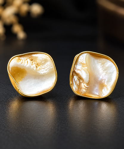 A pair of gold-toned stud earrings with irregular, organic-shaped white mother-of-pearl centers, displayed on a dark textured surface.